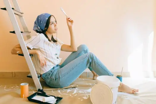 A woman sits barefoot in a room, holding a paintbrush, leaning against a ladder.